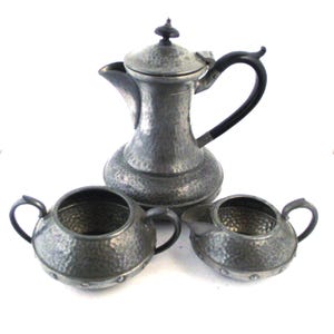 Könnte beinhalten: Ein Vintage-Teeservice aus Zinn mit gehämmerten Texturen. Das Set umfasst eine Teekanne mit schwarzem Griff und Deckel, eine Zuckerdose und einen Sahnekännchen. Die Teile sind auf einer weißen Oberfläche angeordnet.