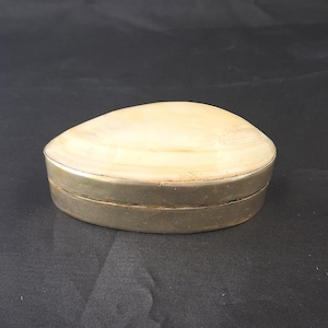 May include: A small, oval-shaped trinket box featuring a smooth, cream-colored stone lid with subtle horizontal banding. The base of the container is constructed from a polished, metallic silver-toned material, creating a clean, simple contrast.