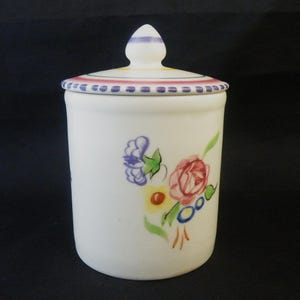 May include: A white ceramic jar with a lid, adorned with a floral design in shades of red, blue, yellow, and green. The lid features a decorative border with pink and purple accents. The jar is a classic kitchenware item.