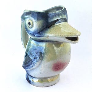 May include: A ceramic jug shaped like a bird with a long beak that serves as the spout. The piece features a glossy glaze in shades of blue, cream, and green, with a small pink patch on the chest and a dark eye detail on the side of the head.