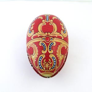 May include: Red egg-shaped decorative box with gold filigree and blue accents. The box features ornate scrollwork and jewel-like embellishments. The design is symmetrical and detailed, with a classic, elegant aesthetic.
