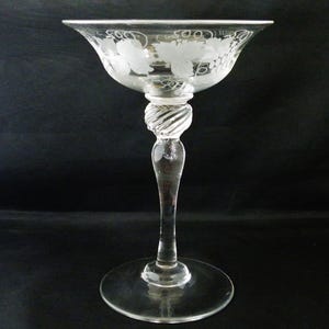 May include: A clear glass goblet with a wide bowl and a long stem. The bowl is decorated with etched grape and leaf designs. The stem features a twisted design. The goblet sits on a round base. The background is black.