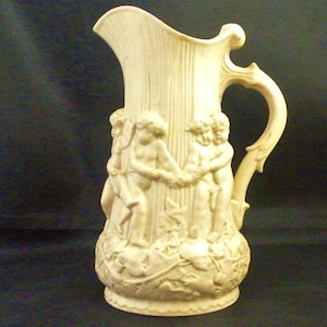 May include: A white ceramic pitcher with a handle and a design of cherubs and vines. The pitcher is decorated with raised details and has a smooth, glossy finish.