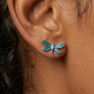 Könnte beinhalten: Nahaufnahme eines türkisblauen Libellen-Ohrsteckers mit einem kleinen Diamanten. Die Libelle hat detaillierte Flügel und einen silberfarbenen Körper. Der Ohrring wird am Ohr einer Person getragen.