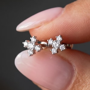 May include: A pair of silver-toned stud earrings, each featuring a cluster of five small, clear, round-cut stones arranged in a cross-like pattern. The metal setting is polished and reflective, holding the stones securely in a symmetrical design.