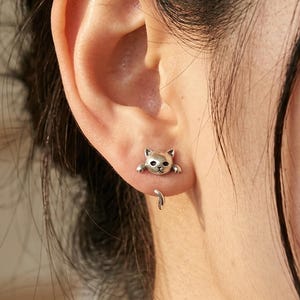 May include: Silver cat-shaped stud earrings. The earrings feature a small, detailed cat design with a curved tail. The cat's head and paws are visible, creating a playful and whimsical look. These earrings are a charming accessory.