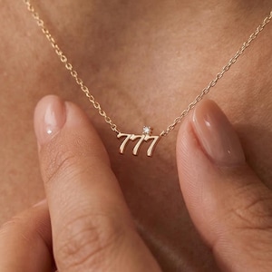 May include: A delicate gold necklace featuring the number "777" with a small gemstone accent. The pendant hangs from a fine chain, creating a minimalist and elegant design. The necklace is shown on a person's neck.
