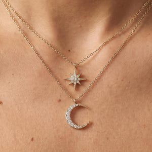 Puede incluir: Un collar dorado de varias capas con un colgante de estrella y un colgante de media luna, ambos adornados con pequeñas piedras brillantes. El collar se muestra en el pecho de una persona.