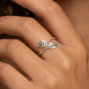 May include: A silver-colored ring featuring a delicate leaf design. The ring wraps around the finger with two detailed leaf accents. The jewelry is shown on a hand, highlighting the ring's elegant and nature-inspired aesthetic.
