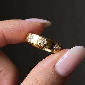 Pode incluir: Um anel de banda em ouro com um padrão repetitivo de motivos florais de quatro pétalas, cada um centrado com uma pequena pedra preciosa redonda e transparente. A superfície metálica é polida e reflexiva, com um perfil liso e arredondado.