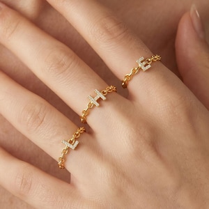 May include: Three gold-toned chain link rings worn on three fingers. Each ring features a single letter charm—L, H, and E—encrusted with small, clear, sparkling stones. The rings are delicate, with a polished metal finish and a flexible chain band.