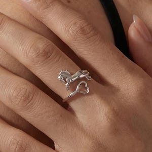 May include: A silver ring with a horse and heart design. The ring features a detailed horse figure and a heart-shaped accent. The ring is worn on a finger.