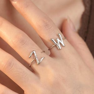 May include: Two silver-toned metal rings worn on the fingers. One ring features the letter Z in a cursive script, and the other ring features the letter W in a cursive script. Both rings have a thin, polished band design that wraps around the finger.