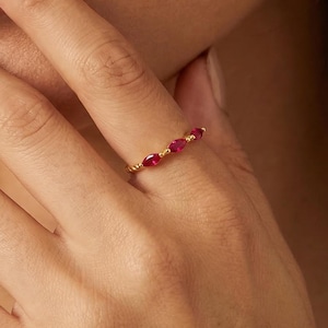 May include: A gold ring with three oval-cut, red gemstones. The ring is on a person's finger, with the gemstones in a row. The ring's band has a textured, twisted design.