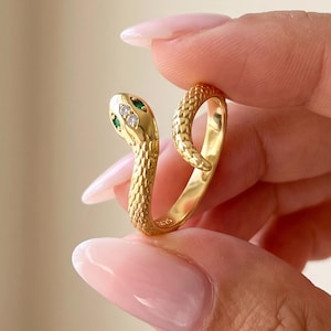 May include: A gold-toned ring shaped like a coiled snake with a textured, scale-like surface. The snake head features two small green stones for eyes and a single clear, sparkling stone set between them on the top of the head.