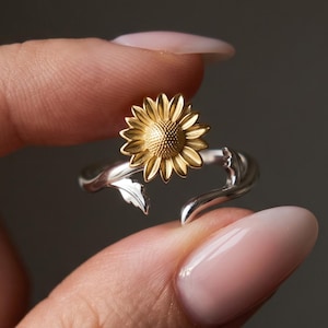 May include: A silver-toned open ring featuring a gold-toned sunflower centrepiece. The band is designed to resemble a stem with a small leaf detail extending from the base of the flower, creating a continuous wrap-around botanical aesthetic.