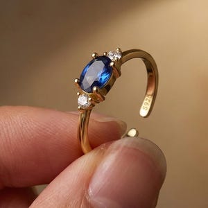 Könnte beinhalten: Ein goldfarbener Ring mit einem großen, ovalförmigen, tiefblauen Edelstein als Mittelpunkt. Zwei kleinere, klare Edelsteine flankieren den Hauptstein. Der Ring ist offen, was eine Größenanpassung ermöglicht. Der Ring wird von einer Person gehalten.