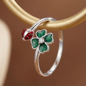Könnte beinhalten: Ein silberner Ring mit einem Marienkäfer- und einem vierblättrigen Kleeblatt-Design. Der Marienkäfer ist rot mit schwarzen Punkten, und das Kleeblatt ist grün mit einem kleinen Edelstein in der Mitte. Der Ring wird vor einem goldfarbenen Hintergrund präsentiert.