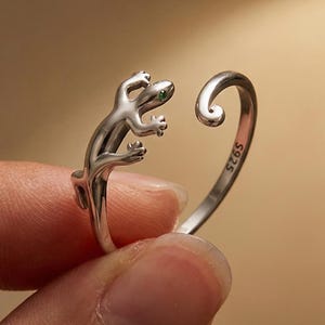 May include: A silver-colored ring featuring a lizard design. The lizard has small, detailed legs and a curved tail. The ring is open-ended, allowing for size adjustment. The lizard's eyes appear to be green. The ring is held by a person's fingers.