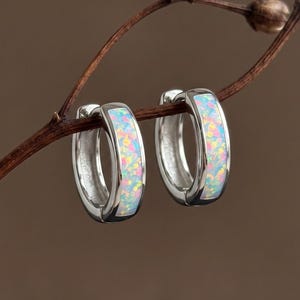 May include: A pair of silver hoop earrings featuring a central inlay of iridescent white opal with flecks of pink, blue, and green color. The earrings are circular in shape with a polished metallic finish and a hinged closure mechanism.