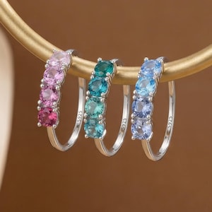 以下が含まれることがあります： カラフルな宝石が並んだ3組のシルバーフープピアス。ピアスはピンクから赤、ティールからターコイズ、明るい青から濃い青へと色のグラデーションを見せています。ピアスは、金色の湾曲したバーに展示されています。