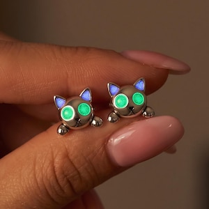 May include: A pair of silver-toned cat-shaped stud earrings featuring glowing green circular eyes and small blue accents on the ears. The cat designs are depicted with paws resting at the bottom, creating a playful and detailed feline appearance.