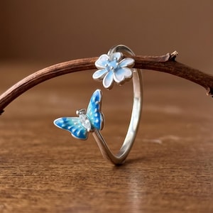 May include: A silver open-style ring featuring a small blue and white enamel butterfly on one end and a blue and white enamel flower on the other. The butterfly has a tiny clear stone at its center, and the ring band is thin and metallic.