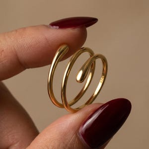 Peut inclure: Une bague bypass dorée présentant un design à double spirale avec des extrémités lisses et arrondies. L'anneau en métal possède une finition polie et comporte un petit poinçon 925 visible sur la surface interne, indiquant une composition en argent.