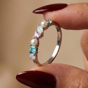 May include: A silver-toned ring featuring a curved arrangement of multi-colored gemstones and pearls. The stones include a square-cut teal gem, a round white pearl, a triangular pink gem, an oval iridescent opal, and a small round white pearl.