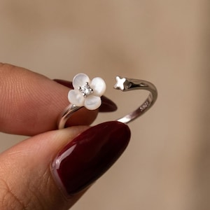 Könnte beinhalten: Ein offener Silberring mit einer kleinen weißen Perlmuttblüte und einem klaren Stein in der Mitte. Die Ringschiene ist mit S925 gestempelt und endet auf der gegenüberliegenden Seite in einem vierzackigen Stern, ein filigranes Schmuckstück.