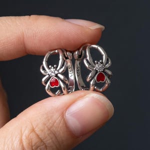 May include: A pair of silver hoop earrings featuring a spider design. Each spider has a small red heart-shaped accent on its abdomen and tiny clear stones on its back. The inner band of the hoop is engraved with the text S925.