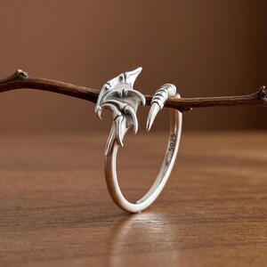 Peut inclure: Une bague en argent présentant un motif d'aile de dragon détaillé à une extrémité et une queue pointue à l'autre. L'anneau est ouvert et comporte un poinçon S925 sur la surface intérieure, indiquant une composition en argent sterling.