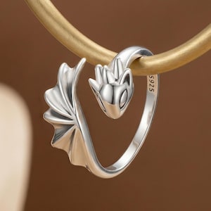 May include: A silver dragon ring with intricate details. The ring features a dragon head and tail design, with a wing on one side. The ring is marked with "S925". The ring is displayed against a neutral background.
