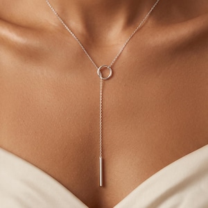 Op de afbeelding: Een zilveren lariat-ketting met een fijne ketting die door een cirkelvormige ring loopt en eindigt in een gladde, cilindervormige staafhanger. De ketting rust op het sleutelbeen en heeft een gepolijste metalen afwerking.