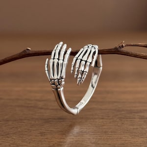 Op de afbeelding: Een zilveren ring met twee skeletachtige handen die om elkaar heen grijpen en in het midden samenkomen. De ring is van gepolijst zilver met donkere, geoxideerde details om de botstructuur te benadrukken. Binnenin staat de tekst S925.