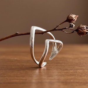 Può includere: Un anello in argento dal design geometrico aperto e angolare appoggiato su un sottile rametto marrone essiccato. La fascia interna dell'anello è chiaramente stampata con il testo S925, che indica la sua composizione in argento sterling e finitura.