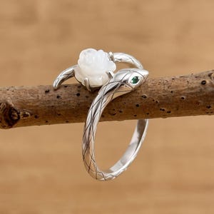 May include: A silver ring shaped like a coiled snake with a textured scale pattern on the band. The snake head features a small green stone for an eye, and a white carved flower is positioned between the snake head and the tail.