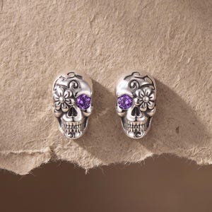 May include: A pair of silver-toned skull earrings featuring intricate floral engravings and a single round purple gemstone set into one eye socket of each skull. The design includes detailed teeth and swirling patterns across the forehead and cheekbones.