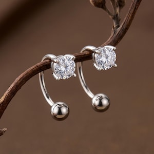 May include: A pair of silver-toned earrings featuring a curved bar design. Each earring has a clear, round-cut gemstone set in a four-prong mount at one end and a smooth, polished metal sphere at the opposite end of the curved metal bar.