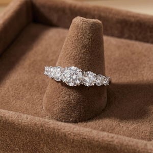 May include: A silver-toned ring featuring five round, clear, faceted stones arranged in a row. The centre stone is the largest, with two smaller stones on each side, all set in a metallic band that reflects light across the clear surfaces.