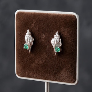May include: A pair of silver stud earrings shaped like spiral seashells, each featuring a small, round, vibrant green gemstone set into the lower portion of the shell structure. The earrings are displayed on a brown, rectangular velvet-like surface.