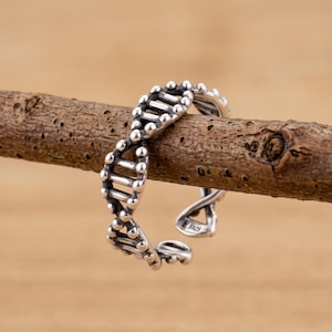 Puede incluir: Un anillo de plata con un diseño de estructura de doble hélice de ADN. La banda está abierta en la parte posterior, lo que permite su ajuste. La superficie metálica tiene un acabado pulido con áreas hundidas oscurecidas y el sello S925.