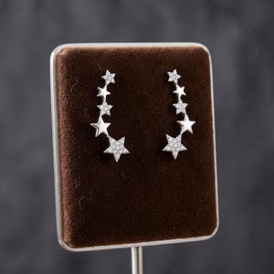 May include: A pair of silver-toned ear climber earrings, each featuring a vertical arrangement of five stars of varying sizes. The stars are encrusted with small, clear, sparkling stones, creating a bright, reflective surface on each piece.