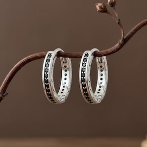 May include: A pair of silver hoop earrings featuring a central row of small, round black stones. The outer edges of the silver hoops are decorated with intricate, engraved scrollwork patterns that wrap around the circular metal frame.