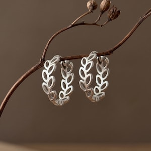 May include: A pair of silver hoop earrings featuring a repeating openwork leaf pattern along the circular band. The earrings are suspended from a thin, dark brown twig with small, dried seed pods attached to the upper branch section.