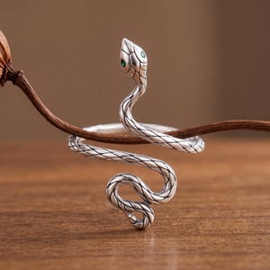 May include: A silver-toned ring shaped like a coiled snake with textured scales and small green eyes. The snake body wraps around to form the band, featuring detailed engravings that mimic natural skin patterns across the entire metal surface.