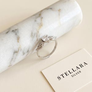 May include: A silver ring with a wing design, featuring a central pear-shaped clear stone and smaller accent stones. The ring is set against a marble background, with a card that reads "STELLARA SILVER".