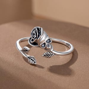 May include: A silver open-style ring featuring a sculpted rose and skull motif on one end and two small leaf details on the other. The band is polished silver with oxidised accents in the crevices and is stamped with the text S925 on the inner side.