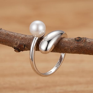May include: A silver open-style ring featuring a single white spherical pearl on one end and a smooth, polished silver teardrop shape on the other. The inner band of the ring is clearly stamped with the text S925, indicating sterling silver.