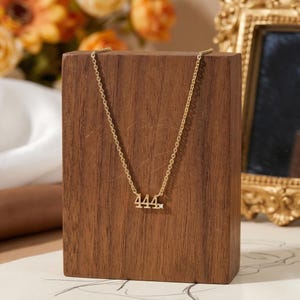 May include: A gold-colored necklace with the number "444" pendant. The necklace is displayed on a wooden block. The background includes a gold-framed mirror and orange flowers.